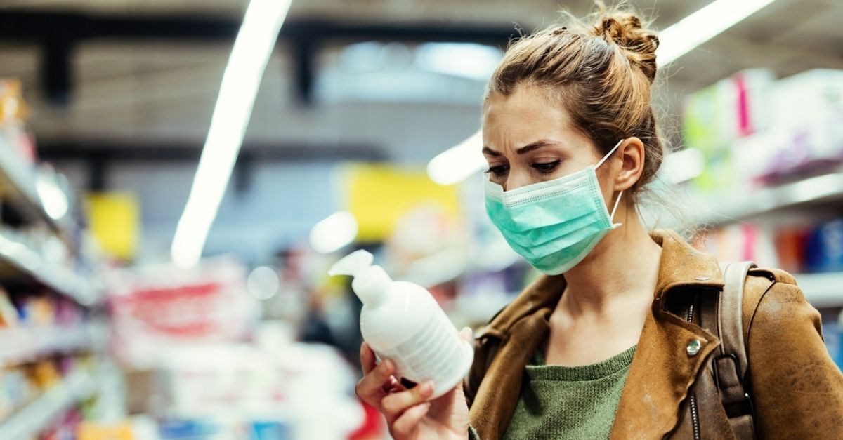 Young woman with protective mask reading label on the bottle while buying hand soap in the supermarket during virus epidemic.